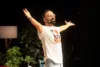Comedian mit Headset-Mikrofon breitet auf der Bühne die Arme aus, im Hintergrund Zimmerpflanzen und Stehlampe